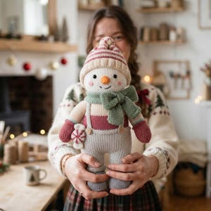 Pode incluir: Um boneco de neve de malha com um gorro às riscas, um cachecol verde sálvia e jardineiras cinzentas com alças. O boneco tem bochechas cor-de-rosa, um nariz cor de laranja e luvas vermelhas, com um pequeno acessório de rebuçado de menta.