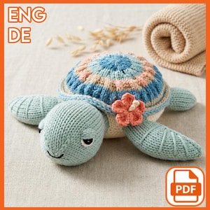 Pode incluir: Uma tartaruga de malha em tons de azul, rosa e creme, com uma flor decorativa. A tartaruga tem uma carapaça e barbatanas texturizadas. A imagem também inclui o texto "ENG DE" e um ícone PDF.