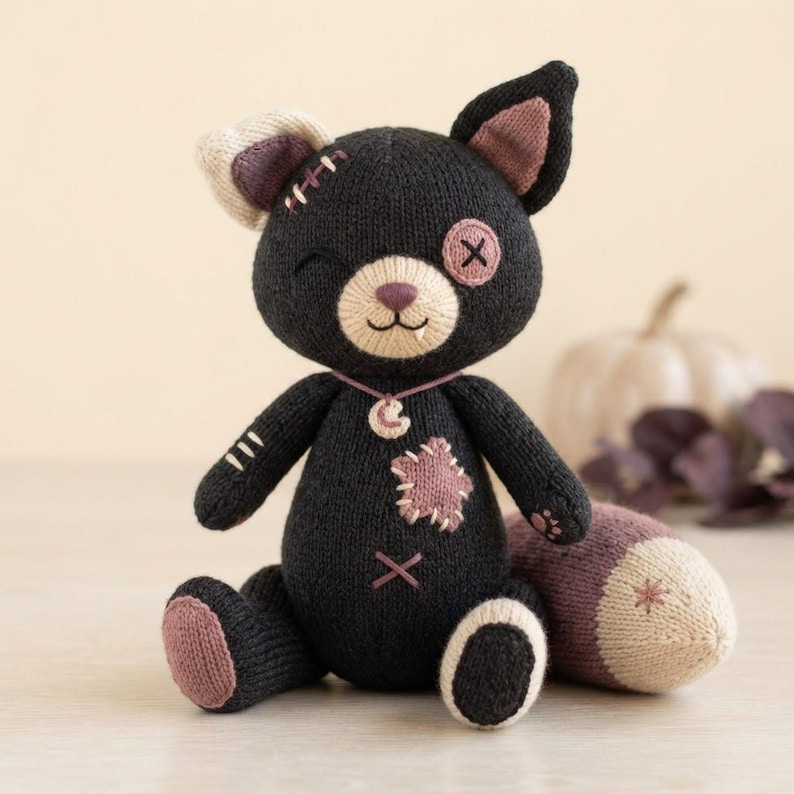 Op de afbeelding: Een zwart gebreid pluche kattenspeeltje met een cr&egrave;mekleurige snuit, &eacute;&eacute;n knoop-oog en &eacute;&eacute;n knipogend oog. Het heeft een roze stervormige patch op de borst, een ketting met een maanhanger en roze accenten op de oren, pootjes en de punt van de staart.
