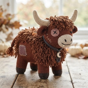 May include: A brown knitted plush toy shaped like a highland cow with shaggy yarn fur, beige horns, and a button eye. It features a dark grey collar with a white crescent moon charm, a light-coloured muzzle, and a small purple patch on its side.