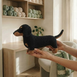 May include: A knitted dachshund toy featuring a black body with tan accents on the muzzle, chest, and paws. The stuffed animal has floppy ears, a long tail, and a textured yarn surface, capturing the distinct elongated shape of the dog breed.