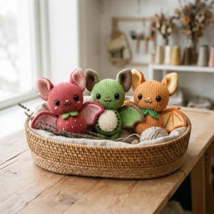 May include: Three small, handmade knitted bat plushies sit inside a woven basket. The left bat is pink with strawberry-like details, the centre bat is green with a cream belly, and the right bat is orange with yellow wing accents and black eyes.