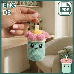 May include: A handmade crochet keychain in the shape of a smiling, light teal cup with a pink and yellow flower on top. The keychain has a gold ring and is held by a person. The image also includes the text "ENG DE" and a PDF icon.