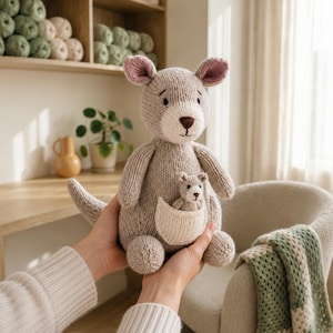 Op de afbeelding: Een handgebreide beige kangoeroe knuffel met een crèmekleurige buidel waarin een kleine bijpassende babykangoeroe zit. De kangoeroe heeft roze gevoerde oren, een donkerbruine geborduurde neus en een lange staart, gemaakt van zacht textielgaren.
