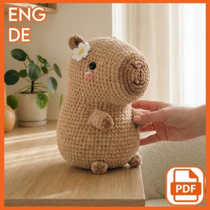 Könnte beinhalten: Ein gehäkeltes Capybara-Plüschtier in Brauntönen, verziert mit einer weißen Blume und rosa Wangen. Das handgefertigte Tier sitzt auf einer Holzoberfläche. Das Bild enthält auch den Text "ENG DE" und ein PDF-Symbol.