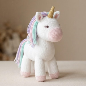 May include: A knitted unicorn toy with a white body, a gold-colored horn, and a pink muzzle. The unicorn features a mane and tail made of pastel-colored yarn strands in shades of light purple, light blue, and pink, with pink-colored hooves.