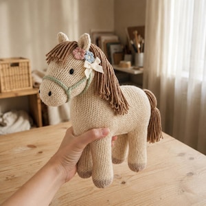 Könnte beinhalten: Ein beiges gestricktes Pferdespielzeug mit brauner Wollmähne und Schweif, einem hellgrünen Halfter und einer kleinen weißen Schleife, die mit zwei winzigen gehäkelten Blumen in Rosa und Blau verziert ist. Das Pferd hat schwarze Augen und dunkle Hufe.