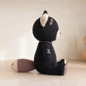 Op de afbeelding: Een gebreid knuffeldier in de vorm van een kat, met een zwart lichaam, cr&egrave;mekleurige binnenkant van de oren en een mauvekleurige staartpunt. Het speelgoed heeft geborduurde stikseldetails op het hoofd en de arm, gesloten ogen en een kleine mauve neus.