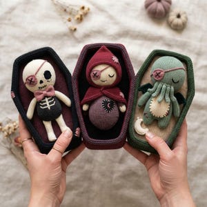 May include: Three crocheted dolls resting in individual coffin-shaped containers. The left doll is a cream-coloured skeleton with a pink bowtie and eyepatch. The centre doll wears a maroon hooded cloak. The right doll is a green creature with tentacles.