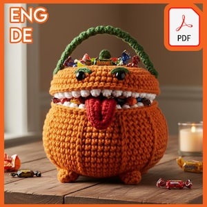 Op de afbeelding: Een oranje gehaakte snoepmand in de vorm van een monster, met een groene handgreep en zwarte ogen. De mand is gevuld met verpakte snoepjes en heeft een rode tong. De mand staat op een houten oppervlak.