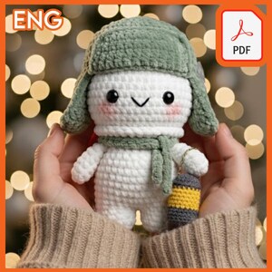 May include: A crocheted snowman doll with a green hat and scarf, holding a small lantern. The snowman has a smiling face with rosy cheeks. The background is blurred with bokeh lights. The image includes a PDF icon in the top right corner.
