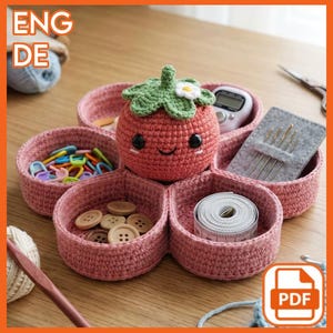Può includere: Un organizer rosa all'uncinetto a forma di fiore, con un pomodoro sorridente al centro. L'organizer contiene forniture per cucito come bottoni, aghi e un metro a nastro. L'immagine include anche il testo "ENG DE" e un'icona PDF.