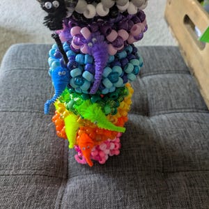 May include: A vibrant stack of beaded creations, resembling fantastical creatures. The stack features a rainbow of colours, including pink, orange, yellow, blue, purple, grey, and white. Each layer has a different creature with pipe cleaner details. The beaded creations are stacked on a grey surface.