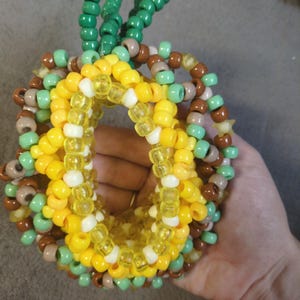 May include: A handmade beaded bracelet shaped like a pineapple. The bracelet is made with yellow, white, green, and brown beads. The pineapple design includes green beaded leaves at the top. The bracelet is held in a hand.