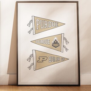 May include: Framed print featuring three pennants with the word "PURDUE" and "BOILER UP" in a serif font. The pennants are gold with black and white accents. The print is in a black frame and leans against a white wall.