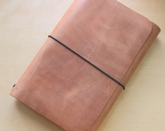 May include: A rectangular notebook cover made of smooth, light brown leather. The cover is secured by a thin, black elastic band that wraps horizontally around the center, holding the folded leather flaps firmly in place against the body.