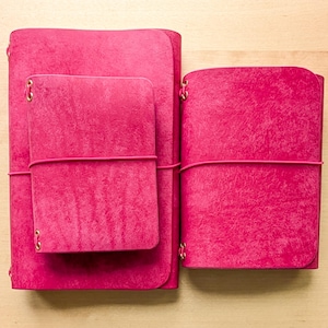 May include: Three bright fuchsia journals with textured covers and rounded corners. The journals are secured with matching elastic bands threaded through metal eyelets on the spines. One large journal sits on the left with a smaller one on top.