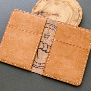 May include: Open, tan leather wallet with white stitching. The wallet has two card slots and a center section with a stamped logo that reads "NO LEATH" and "SINCE 2018". The wallet is open, displaying the interior.