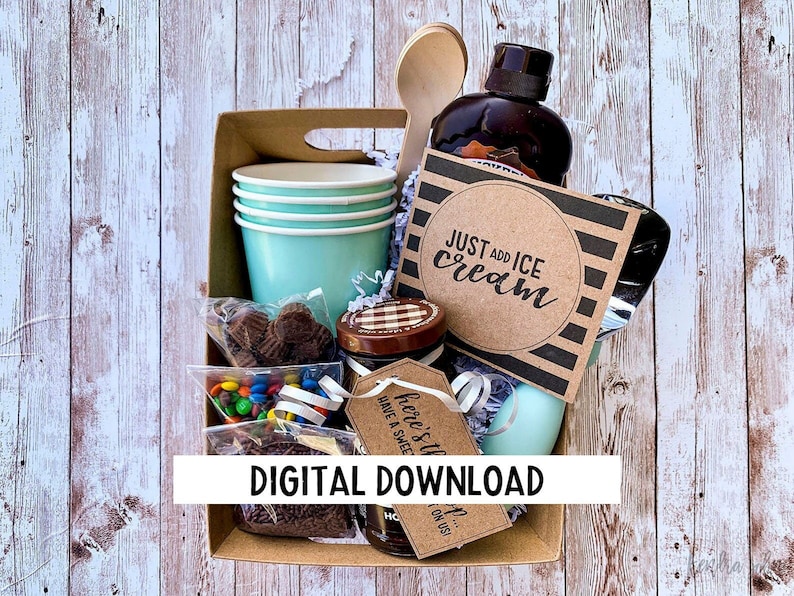 May include: A brown cardboard gift basket filled with a variety of items, including a bottle of chocolate sauce, a wooden spoon, a small jar of chocolate sprinkles, a bag of colourful sweets, a bag of chocolate chips, a stack of light blue paper cups, and a brown card with the text "JUST ADD ICE CREAM".