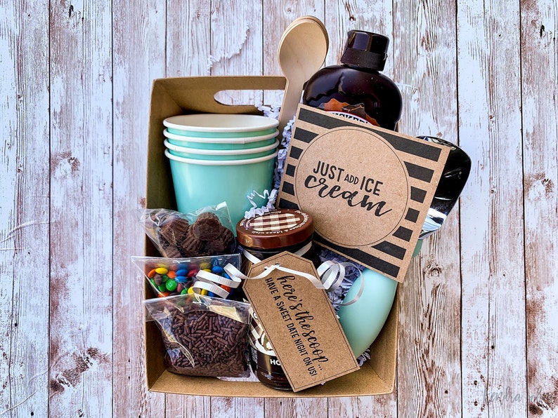 Puede incluir: Una caja de cart&oacute;n marr&oacute;n llena de suministros para helados. La caja contiene una botella de jarabe de chocolate, una cuchara de madera, una bolsa de chispas de chocolate, una bolsa de caramelos de colores, una bolsa de tazas de mantequilla de man&iacute; con chocolate, cuatro vasos de papel azules, una tarjeta a rayas azules y blancas que dice "Just add ice cream", y una tarjeta marr&oacute;n que dice "Here's the scoop. Have a sweet date night on us!"