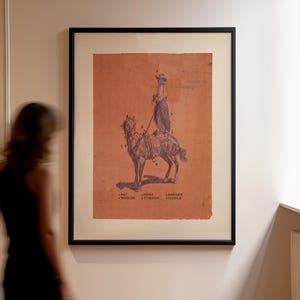 May include: A framed wall art print featuring a dark blue illustration of a person on horseback against a terracotta background. The image includes numbered labels pointing to gear, with text at the bottom listing: 1 HAT, 2 WOOLIES, 3 SPURS, 4 STIRRUPS, 5 HARNESS, 6 SADDLE.