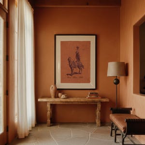 May include: A framed print featuring a dark illustration of a person riding a camel on a terracotta-coloured background, displayed above a long wooden console table with two ceramic vessels and a stack of books, next to a floor lamp and bench.