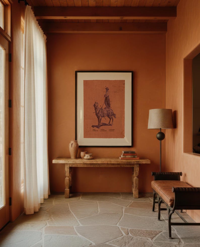 May include: A framed print featuring a dark illustration of a person riding a camel on a terracotta-coloured background, displayed above a long wooden console table with two ceramic vessels and a stack of books, next to a floor lamp and bench.