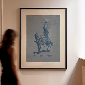 May include: A framed wall art print featuring a blue-toned illustration of a person on horseback. The image includes numbered labels pointing to specific gear, with text at the bottom listing: 1 HAT, 2 WOOLIES, 3 SPURS, 4 STIRRUPS, 5 HARNESS, 6 SADDLE.