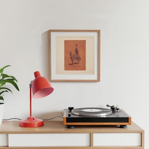 May include: A square framed print featuring a dark brown illustration of a person riding a horse on a terracotta-coloured background. The artwork is displayed in a light wood frame with a white mat, positioned above a black and wood record player.