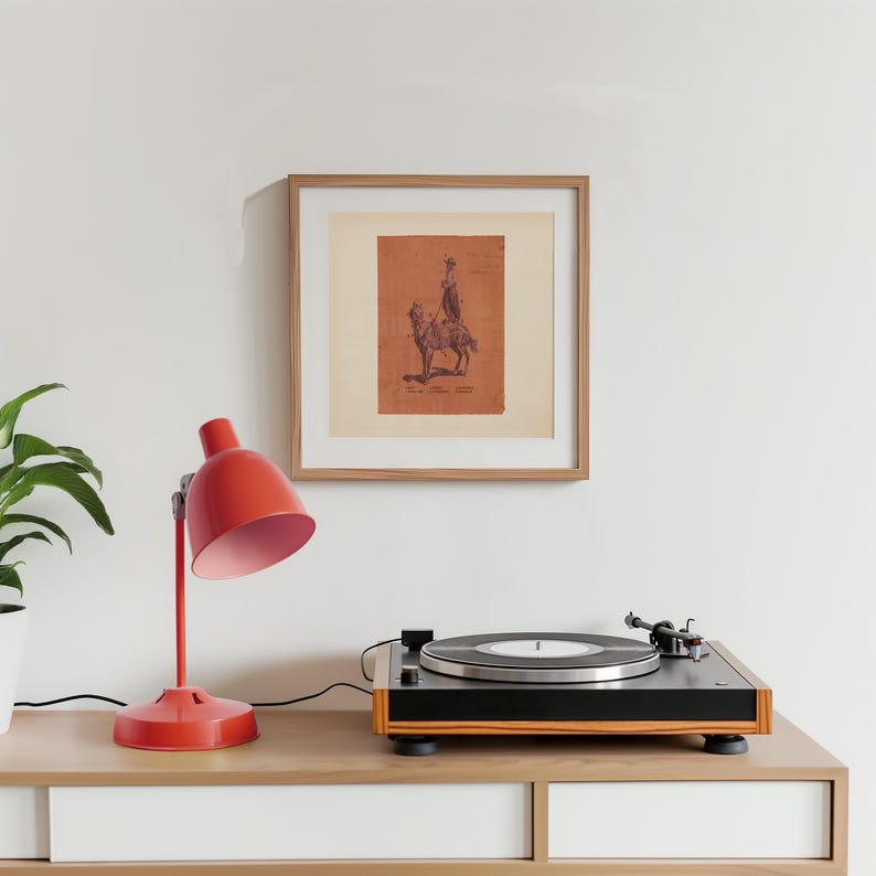 May include: A square framed print featuring a dark brown illustration of a person riding a horse on a terracotta-coloured background. The artwork is displayed in a light wood frame with a white mat, positioned above a black and wood record player.