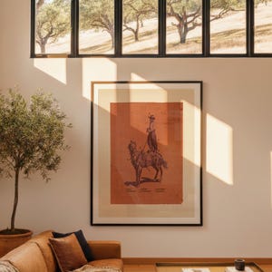 May include: A framed wall art print featuring a dark illustration of a person riding a horse on a burnt orange background. The print is displayed in a black frame on a light-coloured wall, positioned above a tan leather sofa and a gold coffee table.