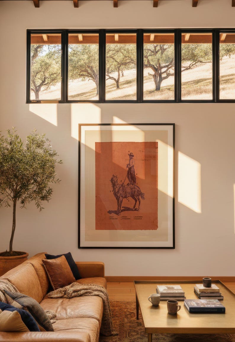 May include: A framed wall art print featuring a dark illustration of a person riding a horse on a burnt orange background. The print is displayed in a black frame on a light-coloured wall, positioned above a tan leather sofa and a gold coffee table.