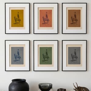 May include: Six framed prints arranged in two rows, each featuring a dark illustration of a person riding a camel. The prints have varied background colours including mustard, terracotta, brown, blue, green, and grey, all within black frames.