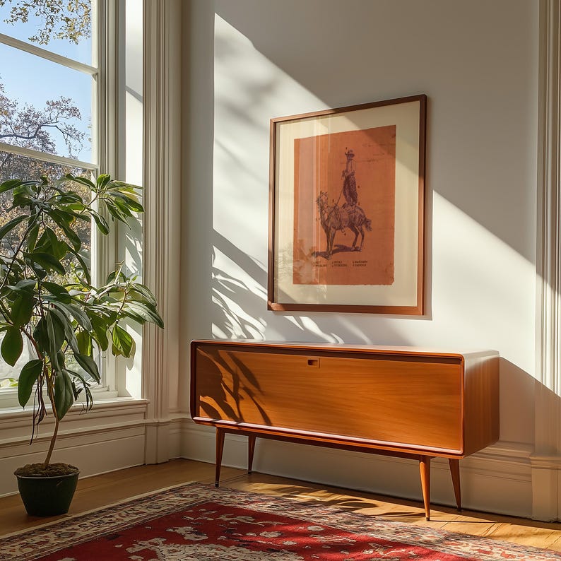 May include: A rectangular wooden sideboard with a smooth finish and four tapered legs, positioned below a framed print featuring a monochromatic illustration of a person riding a llama, with text printed at the bottom of the artwork.