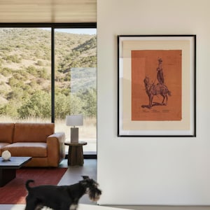 May include: A framed wall art print featuring a dark red illustration of a person riding a horse on a light beige background. The artwork includes small text labels near the figure and horse, all enclosed within a simple black frame on a white wall.