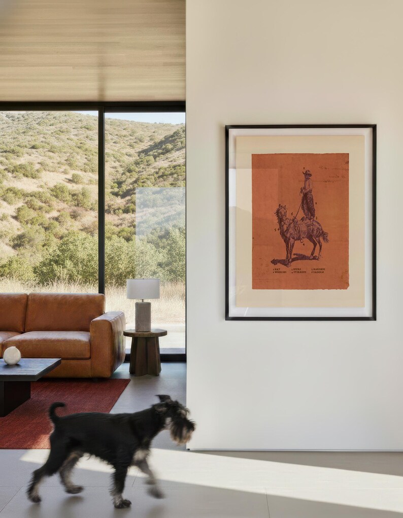 May include: A framed wall art print featuring a dark red illustration of a person riding a horse on a light beige background. The artwork includes small text labels near the figure and horse, all enclosed within a simple black frame on a white wall.