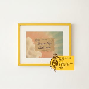 May include: A framed print with a yellow frame. The print features a vintage postcard design with the text "Dream big, little one." The background shows a sky with clouds. A yellow banner with text and a cowboy graphic is attached to the frame.