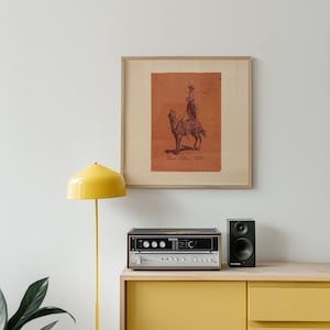 May include: A framed square print featuring a dark brown ink illustration of a person riding a camel on a terracotta orange background. The print includes the text "LEAF", "LIVING", and "LANDSCAPE" printed in small black letters at the bottom.