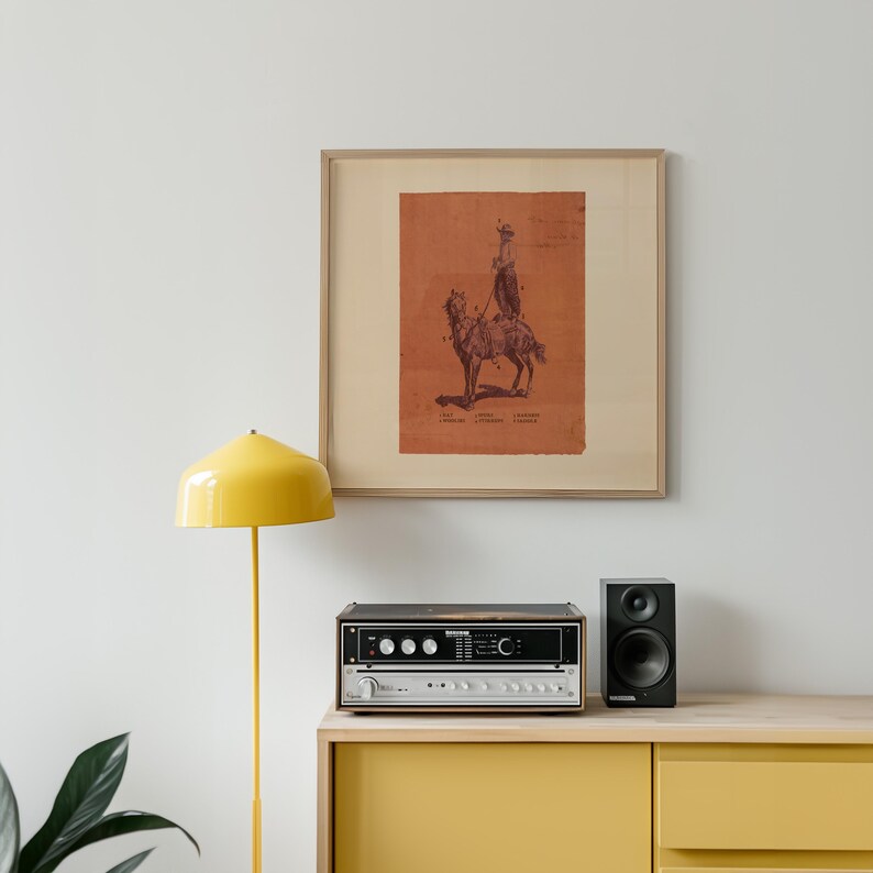 May include: A framed square print featuring a dark brown ink illustration of a person riding a camel on a terracotta orange background. The print includes the text "LEAF", "LIVING", and "LANDSCAPE" printed in small black letters at the bottom.