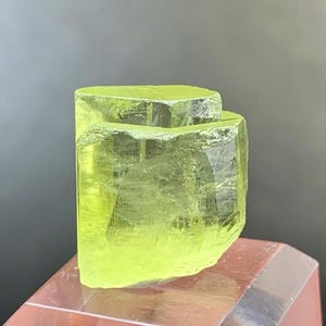 以下が含まれることがあります： プリズム状の幾何学的な構造と内部の亀裂が見える、半透明のシャルトルーズ色の結晶標本。この鉱物は鋭く平らなファセットと鮮やかなネオンイエローの色合いが特徴で、透明な多面体の台座の上に置かれています。