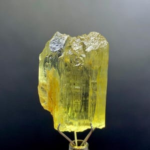 Könnte beinhalten: Ein durchscheinendes, gelb-grünes Kristallexemplar. Das grob behauene Mineral wird von einer Metallklemme vor einem dunklen Hintergrund gehalten. Die Oberfläche des Kristalls hat ein strukturiertes, gestreiftes Aussehen mit Farb- und Opazitätsschwankungen.