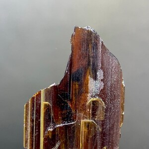 Puede incluir: Primer plano de un espécimen mineral marrón translúcido con estrías verticales y brillo metálico. El mineral está montado sobre un soporte metálico plateado, mostrando su forma natural y en bruto. El color varía de marrón oscuro a ámbar.