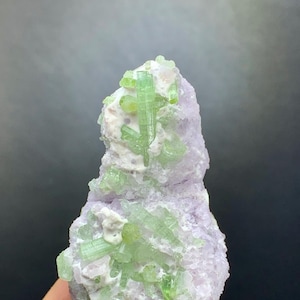 Peut inclure: Spécimen minéral naturel avec des cristaux de tourmaline vert clair incrustés dans une matrice de pierre violet pâle et blanche. Les cristaux sont allongés et ont un aspect vitreux.
