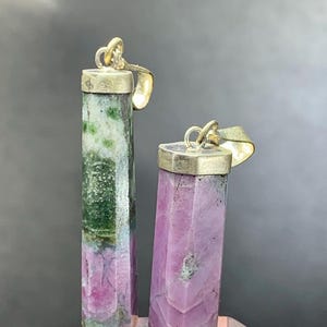Könnte beinhalten: Zwei Edelsteinanhänger. Einer ist ein hoher, rechteckiger Anhänger mit einem grün-rosa Farbschema. Der andere ist ein kürzerer, rechteckiger Anhänger mit einem rosa Farbschema. Beide Anhänger haben eine silberfarbene Kappe und Schlaufe.
