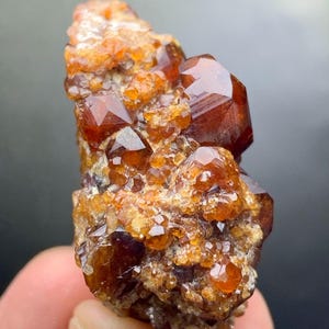 May include: A cluster of orange-brown garnet crystals. The crystals have a glossy, faceted appearance, with a mix of small and large formations. The overall structure is rough and textured, with a natural, earthy aesthetic. The mineral is held in a hand.