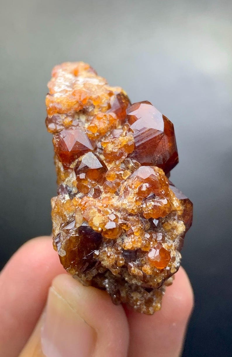 May include: A cluster of orange-brown garnet crystals. The crystals have a glossy, faceted appearance, with a mix of small and large formations. The overall structure is rough and textured, with a natural, earthy aesthetic. The mineral is held in a hand.