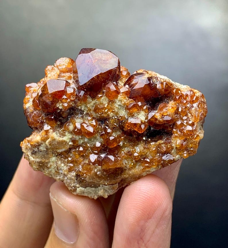 May include: A cluster of lustrous, reddish-brown garnet crystals on a matrix of tan rock. The crystals have a faceted, geometric shape, reflecting light. The overall appearance is rough and natural, showcasing the beauty of the mineral formation.