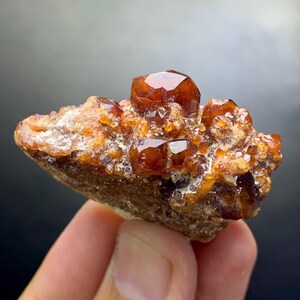 May include: A cluster of vibrant, orange-brown crystals, possibly garnet, sits atop a rough, earthy matrix. The crystals have a faceted, gem-like appearance, catching the light. The overall color palette is warm, with shades of brown and orange.