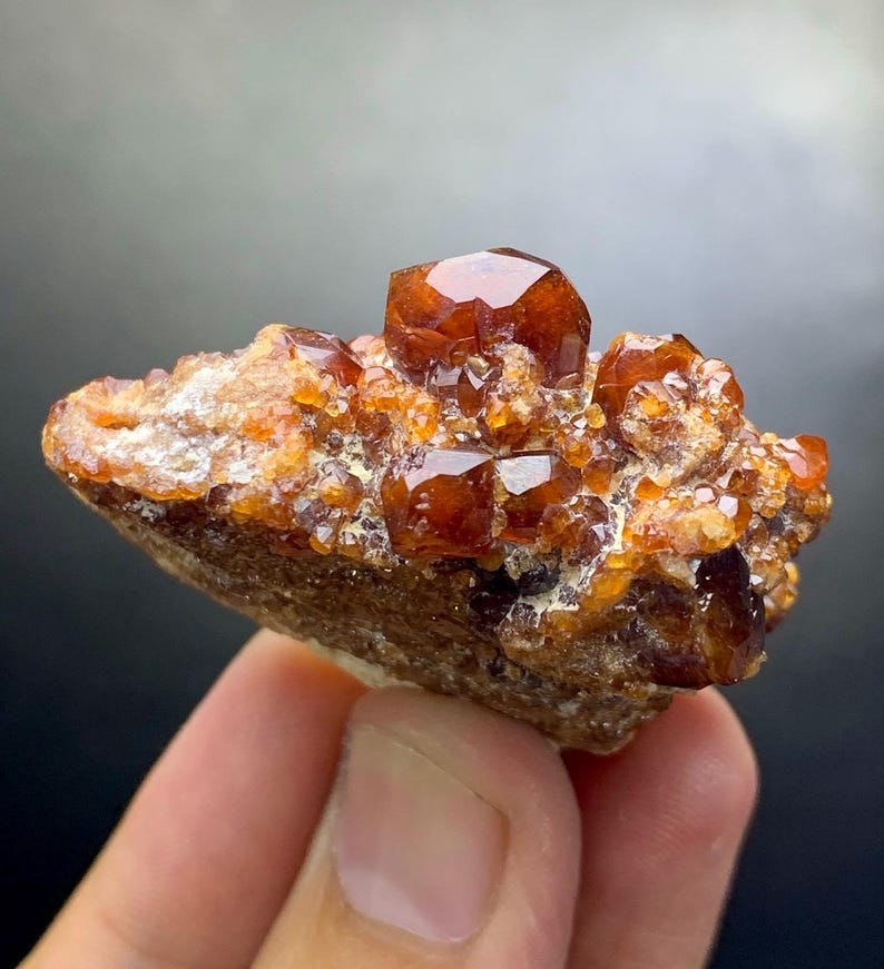 May include: A cluster of vibrant, orange-brown crystals, possibly garnet, sits atop a rough, earthy matrix. The crystals have a faceted, gem-like appearance, catching the light. The overall color palette is warm, with shades of brown and orange.