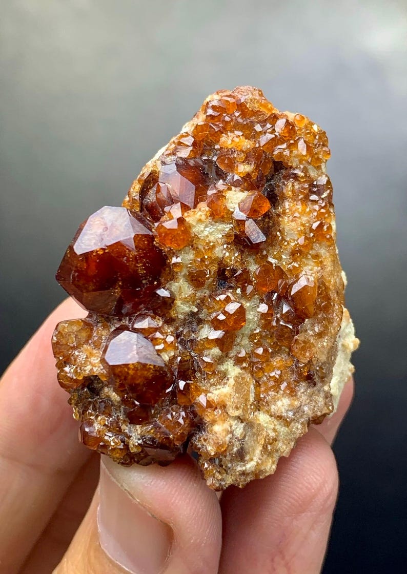 May include: A cluster of lustrous, reddish-brown garnet crystals on a matrix of tan rock. The crystals have a glassy appearance and are of varying sizes, with some displaying well-defined geometric shapes. The overall texture is rough and natural.
